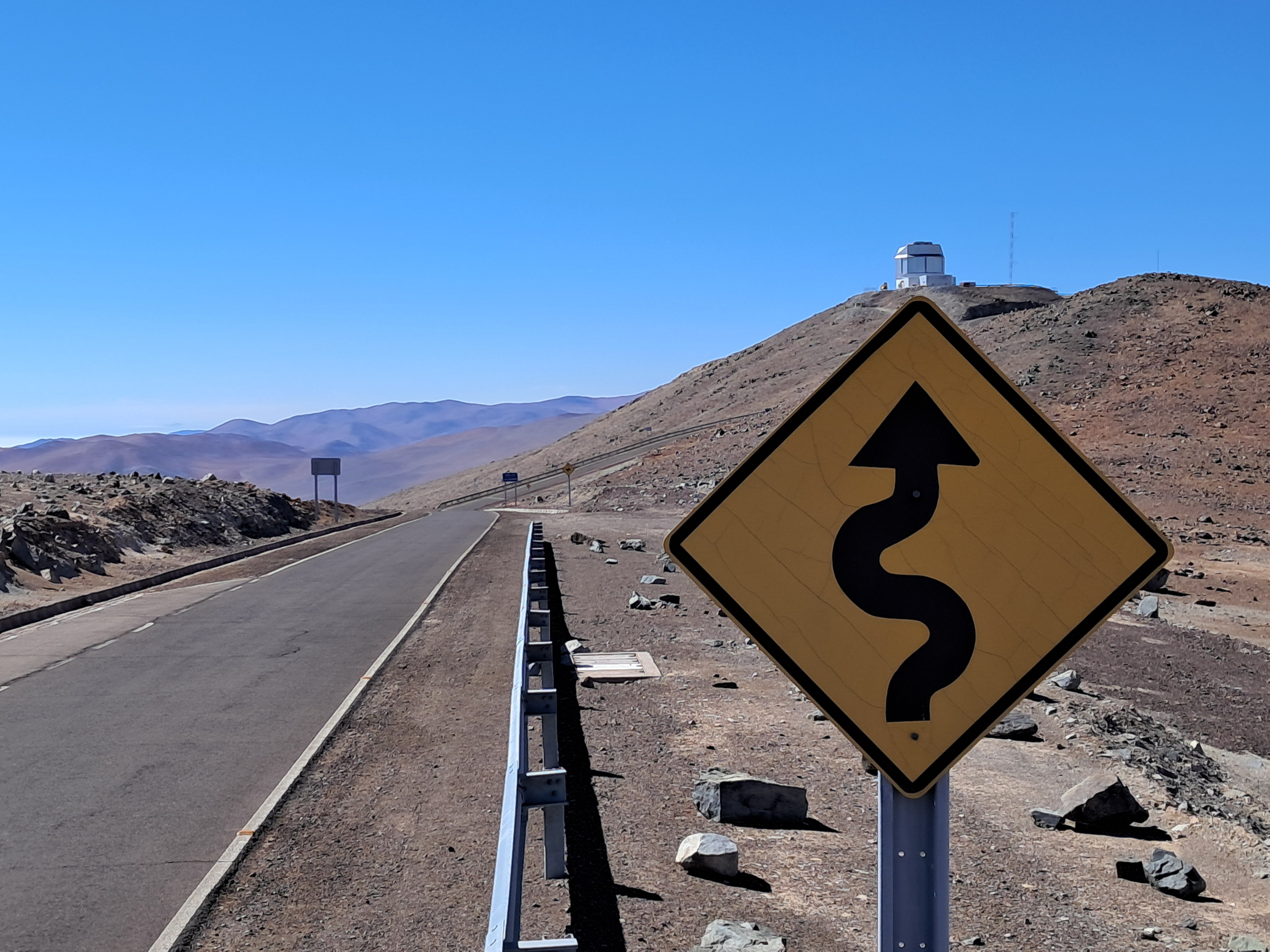 A yellow road sign showing a winding arrow standing on a desert mountain road leaning towards the 4MOST site, symbolising the twists and turns of the journey to first light.
